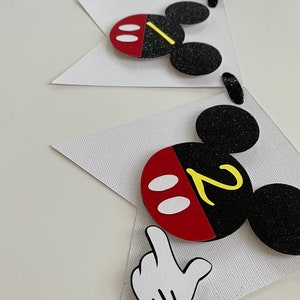 以下が含まれることがあります： 白いバナーに、黒と赤のミッキーマウスの頭が、各旗に黄色の数字2が書かれています。ミッキーマウスの頭は耳にキラキラしていて、白い手袋が下を向いています。