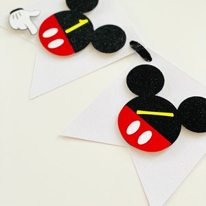 以下が含まれることがあります： 黒と赤のミッキーマウスの頭が付いた白いバナー。バナーには黒い紐が付いています。ミッキーマウスの頭には黄色のアクセントと白い目の円があります。