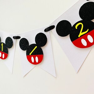 以下が含まれることがあります： 黒い耳、赤と白の円、黄色の数字1と2が付いたミッキーマウスをテーマにしたバナー。バナーは白い厚紙で作られており、黒い紐で繋げられています。