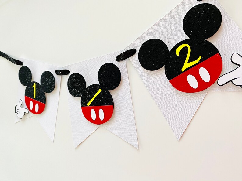 以下が含まれることがあります： 黒い耳、赤と白の円、黄色の数字1と2が付いたミッキーマウスをテーマにしたバナー。バナーは白い厚紙で作られており、黒い紐で繋げられています。