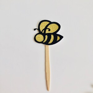 May include: A decorative bee-shaped cupcake topper. The bee has a gold glitter body and black outline. It is attached to a wooden stick. The topper is designed for use in baked goods or other decorative purposes.