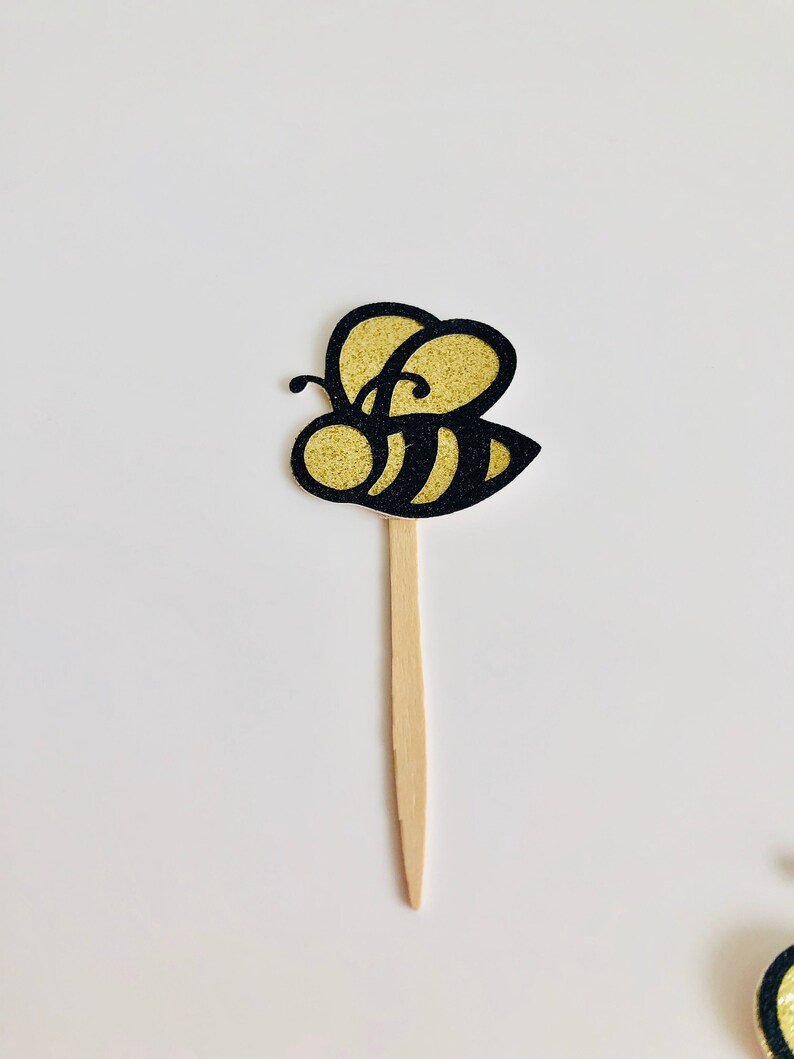 May include: A decorative bee-shaped cupcake topper. The bee has a gold glitter body and black outline. It is attached to a wooden stick. The topper is designed for use in baked goods or other decorative purposes.