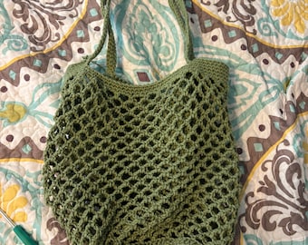 May include: A handmade green crochet tote bag with a net-like design. The bag has two handles and a solid base. A pair of black scissors and a green crochet hook are visible in the background.