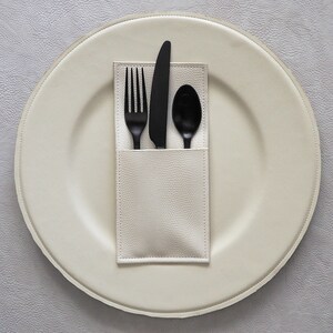 May include: A white leather placemat with a white napkin holder containing a black fork, knife, and spoon.