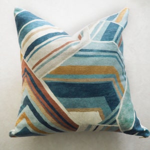 May include: A decorative pillow with a geometric pattern in shades of blue, brown, and gold. The pillow has a white background and features a modern, abstract design.