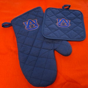 May include: A set of two navy blue quilted kitchen accessories featuring an orange and blue embroidered "AU" logo. The set includes a pot holder and an oven mitt.
