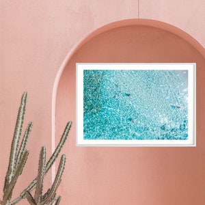 May include: A white metal chair with a curved back and a white framed picture of a turquoise blue ocean with white foam. The chair and picture are in front of a pink wall.