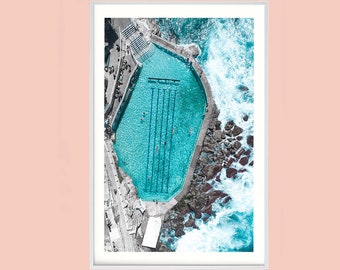 May include: An aerial view of an oceanfront pool with turquoise water and white tiled lanes. The pool is surrounded by rocks and crashing waves.