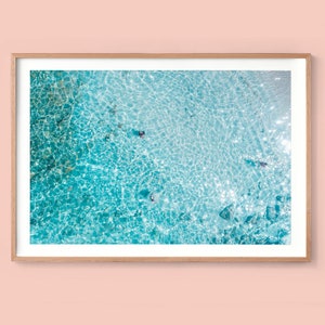 May include: An aerial view of turquoise blue water with ripples and sunlight reflecting on the surface.  Several people are swimming in the water.