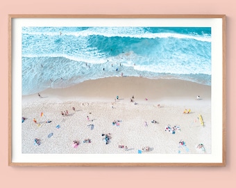 May include: Aerial view of a beach scene with turquoise waves crashing on the shore. People are scattered on the sandy beach, some sunbathing, others near the water. The image is framed in a light brown wooden frame.