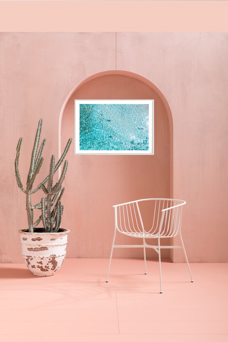 May include: A white metal chair with a curved back and a white framed picture of a turquoise blue ocean with white foam. The chair and picture are in front of a pink wall.