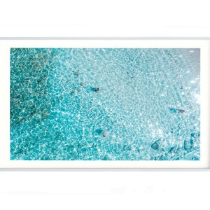 May include: An aerial view of a turquoise blue ocean with ripples and waves. The water is clear and shallow, revealing the sandy bottom.