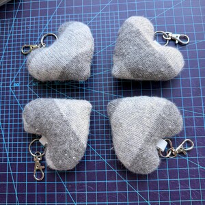 May include: Four heart-shaped keyrings made of grey and white knit fabric. Each plush heart features a silver-toned metal lobster clasp attached to a split ring, secured by a small white fabric loop at the top of the heart-shaped textile form.