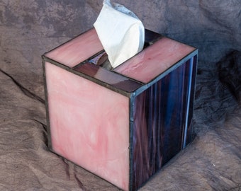 May include: A pink and purple stained glass tissue box holder with a white tissue peeking out of the top.