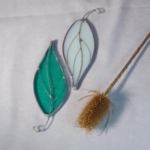 May include: Two stained glass leaf ornaments, one teal and one white, with silver-colored wire detailing. The leaves have a delicate, veined design and a loop at the top for hanging. A dried thistle adds a natural element to the composition.