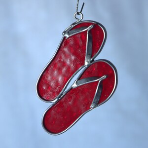 May include: A pair of red stained glass flip-flops with silver-colored metal edging and straps. The flip-flops are hanging from a silver-colored metal hook. The glass has a textured appearance.