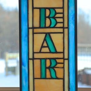 May include: A stained glass window hanging in a window frame. The window is rectangular and features the word "BAR" in green letters on a yellow background. The letters are outlined in black and the background is surrounded by a blue border with red accents.