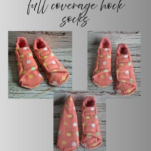 以下が含まれることがあります： 白、黄、緑のドット柄のピーチ色のハックソックス。靴下は柔らかいピンク色の素材で裏打ちされ、ベルクロ留めが付いています。「full coverage hock socks」というテキストが筆記体で書かれています。