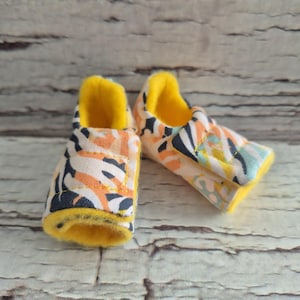 以下が含まれることがあります： オレンジ、ネイビー、ライトブルーのカラフルな花柄のベビーシューズのペア。内側とトリムは明るい黄色です。靴は柔らかい生地で作られています。