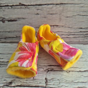 以下が含まれることがあります： ピンク、黄色、白の花柄のベビーシューズのペア。内側とトリムは黄色です。靴は布製で、柔らかく、乳児に適しているように見えます。背景は明るい木です。