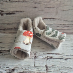 以下が含まれることがあります： 柔らかいクリーム色の内側と明るい灰色の外側を備えた2つのベビーブーティー。片方のブーティーには赤と白のキノコのプリント、もう片方には緑の葉の模様があります。ブーティーは快適さと暖かさを考慮して設計されています。