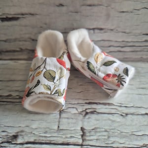 以下が含まれることがあります： 花柄のベビーブーティーのペア。白い生地には、赤、緑、黄色の花と葉が飾られています。ブーティーの内側は、柔らかいクリーム色の素材です。