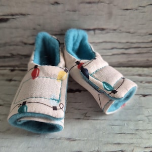 以下が含まれることがあります： カラフルなクリスマスライトのイラストが施された白い生地のアウトドアベビーブーティーのペア。内側は柔らかい水色の生地で裏打ちされています。ブーティーは、シンプルなスリップオンスタイルでデザインされています。