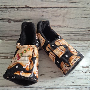 以下が含まれることがあります： ジンジャーブレッドマン柄のベビーブーティーのペア。ブーティーは黒い内側とトリムで、黒い背景に茶色とベージュのジンジャーブレッドマンのデザインが施されています。柔らかい生地で作られたブーティーです。