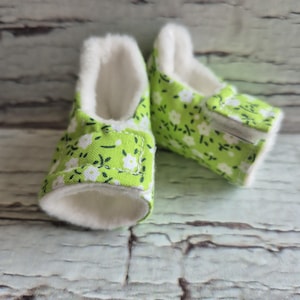 以下が含まれることがあります： 緑色の花柄と白いフェイクファーの裏地が付いたベビーシューズのペア。靴は布製で、柔らかくクッション性のある内側があります。靴は乳児や幼児向けに設計されています。
