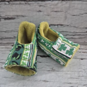 以下が含まれることがあります： 緑と白のクローバー模様のベビーシューズのペア。靴は柔らかい黄色の内側とベルクロ留めです。乳幼児向けに設計された靴です。