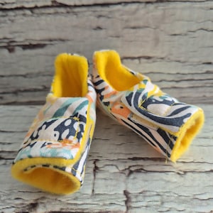 以下が含まれることがあります： カラフルな花柄とゼブラストライプ模様のベビーシューズのペア。靴の内側とトリムは黄色です。靴は布製で、ベルクロ留めです。