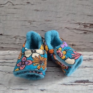 以下が含まれることがあります： 花柄のベビーブーティーのペア。ブーティーは、オレンジ、ピンク、白の花が咲くターコイズブルーの背景を持っています。ブーティーの内側は、柔らかい水色の素材です。