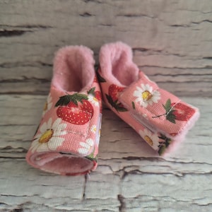 以下が含まれることがあります： ピンクのイチゴとデイジー柄のベビーブーティーのペア。内側は柔らかなピンク色のフェイクファーで裏打ちされています。ブーティーは明るい木目調の背景に配置されています。