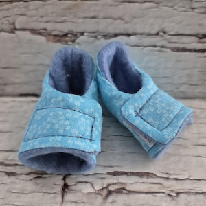以下が含まれることがあります： 白の花柄が施された、水色の生地で作られたベビーブーティーのペア。内側は濃い青色で、ブーティーは面ファスナーで留められています。背景は明るい茶色の風化した木材です。