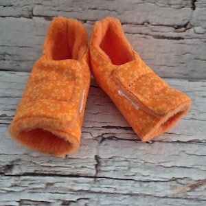 以下が含まれることがあります： オレンジ色のベビーブーティーのペア。ブーティーは、花柄の柔らかいキルト生地で作られています。内側は明るいオレンジ色で、ブーティーにはベルクロ留めが付いています。