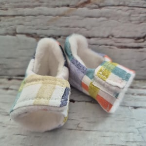 以下が含まれることがあります： 黄色、オレンジ、青、白のカラフルなチェック柄のベビーシューズのペア。靴は柔らかい白い内側とベルクロストラップが付いています。乳児向けに設計された靴です。