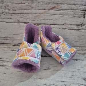 May include: A pair of baby booties with a colorful geometric pattern in orange, yellow, blue, and purple. The interior is lined with soft purple fabric. These handmade booties are designed for comfort and style.