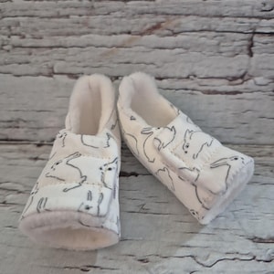 以下が含まれることがあります： ベビーブーティーのペア。白い生地には、ウサギの黒い線画が描かれています。内側とトリムは柔らかく、明るいベージュです。ブーティーは面ファスナーで留められています。