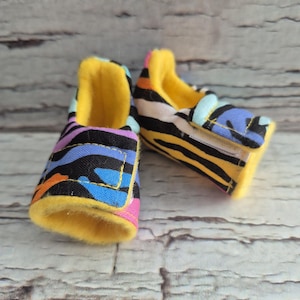 以下が含まれることがあります： カラフルなゼブラ柄のデザインのベビーシューズのペア。靴は、黄色、青、オレンジ、ピンクのストライプが入った黒いベースが特徴です。内側とトリムは明るい黄色です。