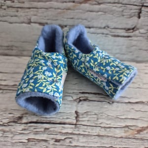 以下が含まれることがあります： 青い花柄が施された、ターコイズ色の背景のベビーブーティーのペア。内側は柔らかい水色の素材です。ブーティーは、明るい茶色の木製の背景に置かれています。