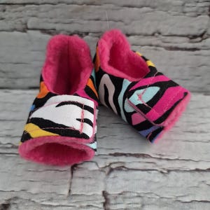 以下が含まれることがあります： カラフルなゼブラ柄のデザインのベビーシューズのペア。靴はピンク色のフェイクファーの裏地と柔らかく柔軟なソールを備えています。外側は黒、白、ピンク、黄色、青の模様が混ざっています。