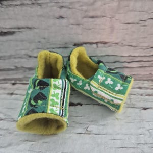 May include: A pair of baby shoes with a green shamrock pattern. The shoes have a yellow interior and a white and black stripe design. The shoes are made of fabric and are designed for infants.