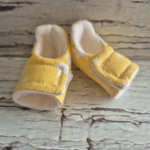 以下が含まれることがあります： 白い星模様の黄色いベビーシューズのペア。靴は柔らかい白い内側とベルクロストラップ留めが付いています。靴は柔らかい生地で作られており、快適さを追求したデザインです。