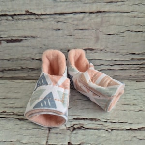 以下が含まれることがあります： グレー、ピーチ、白の色合いの幾何学模様が特徴のベビーブーティーのペア。内側はソフトピーチ色です。これらの小さな靴は乳児向けに設計されており、柔らかい生地で作られています。