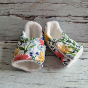 以下が含まれることがあります： 花柄のベビーブーティーのペア。生地は、赤、黄、青、緑のカラフルな花が咲く白い背景が特徴です。ブーティーの内側は、白くふわふわした素材で裏打ちされています。