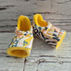 以下が含まれることがあります： 白地を背景に、黄色、オレンジ、ターコイズのフローラル柄のベビーシューズのペア。内側とトリムは明るい黄色です。靴は、風化した明るい色の木の背景に置かれています。