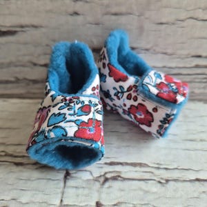 以下が含まれることがあります： 白地に赤、青、紫の花柄のベビーブーティーのペア。内側とトリムは鮮やかな青色です。ブーティーは柔らかい生地でできており、快適に見えます。