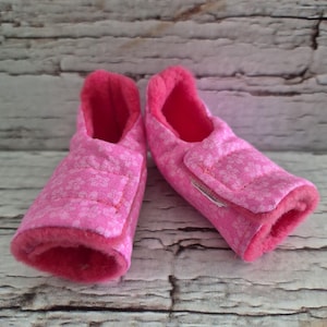 May include: Pair of pink baby booties with a floral pattern. The booties have a soft, fuzzy interior and a velcro strap closure. The exterior fabric is a light pink with a repeating floral design.