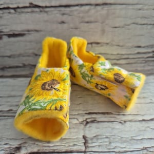 以下が含まれることがあります： 明るい黄色の内側と白い背景にヒマワリ模様が施されたベビーシューズ。 柔らかい生地で作られた靴は快適さを追求したデザインです。 背景は明るい木目です。