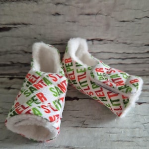 以下が含まれることがあります： 白、赤、緑の模様が入ったベビー用スリッパのペア。「Santa's Little Helper」の文字が入っています。スリッパの内側は白くてふわふわです。